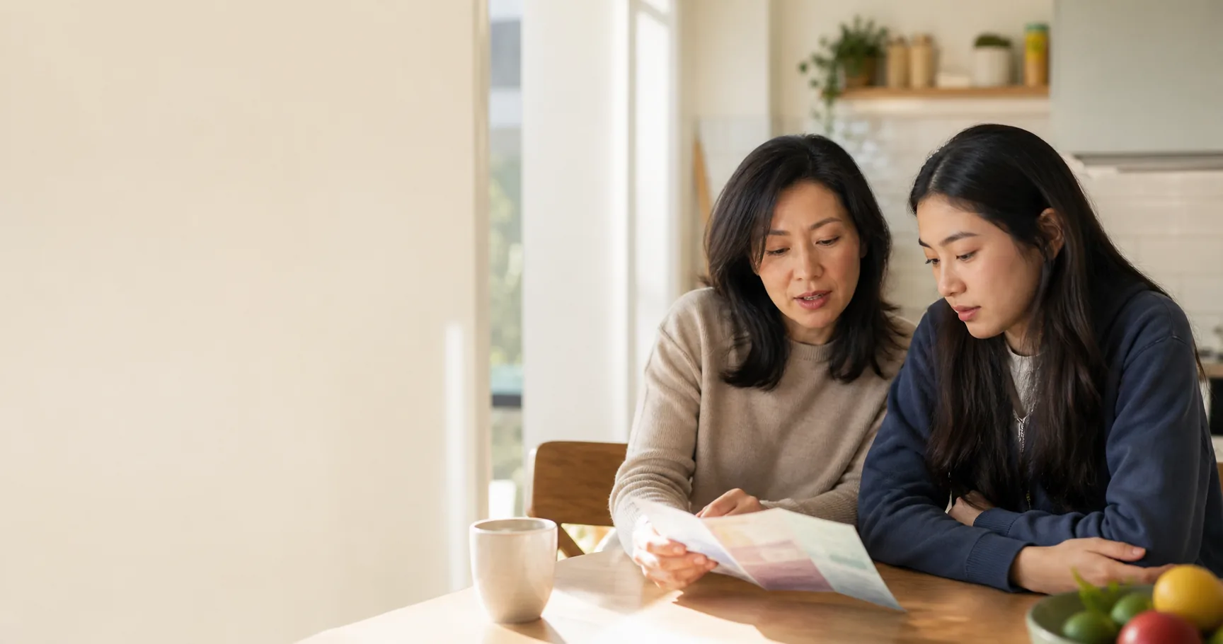 An East Asian mother and older teenage daughter reading health information together in a bright kitchen